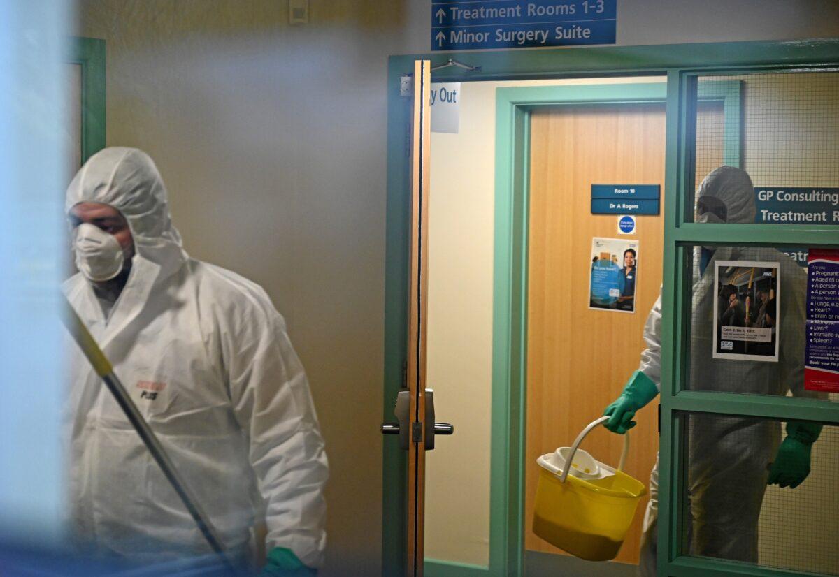Workers dressed in protective clothing, including face masks and gloves, are pictured carrying buckets as they work inside of at the Warmdene doctor's Surgery at County Oak Medical Centre in Brighton, southern England on Feb. 10, 2020, after it closed for "urgent operational health and safety reasons," following reports a member of staff was infected with the 2019-nCoV strain of the novel coronavirus. (Glyn Kirk//AFP via Getty Images)