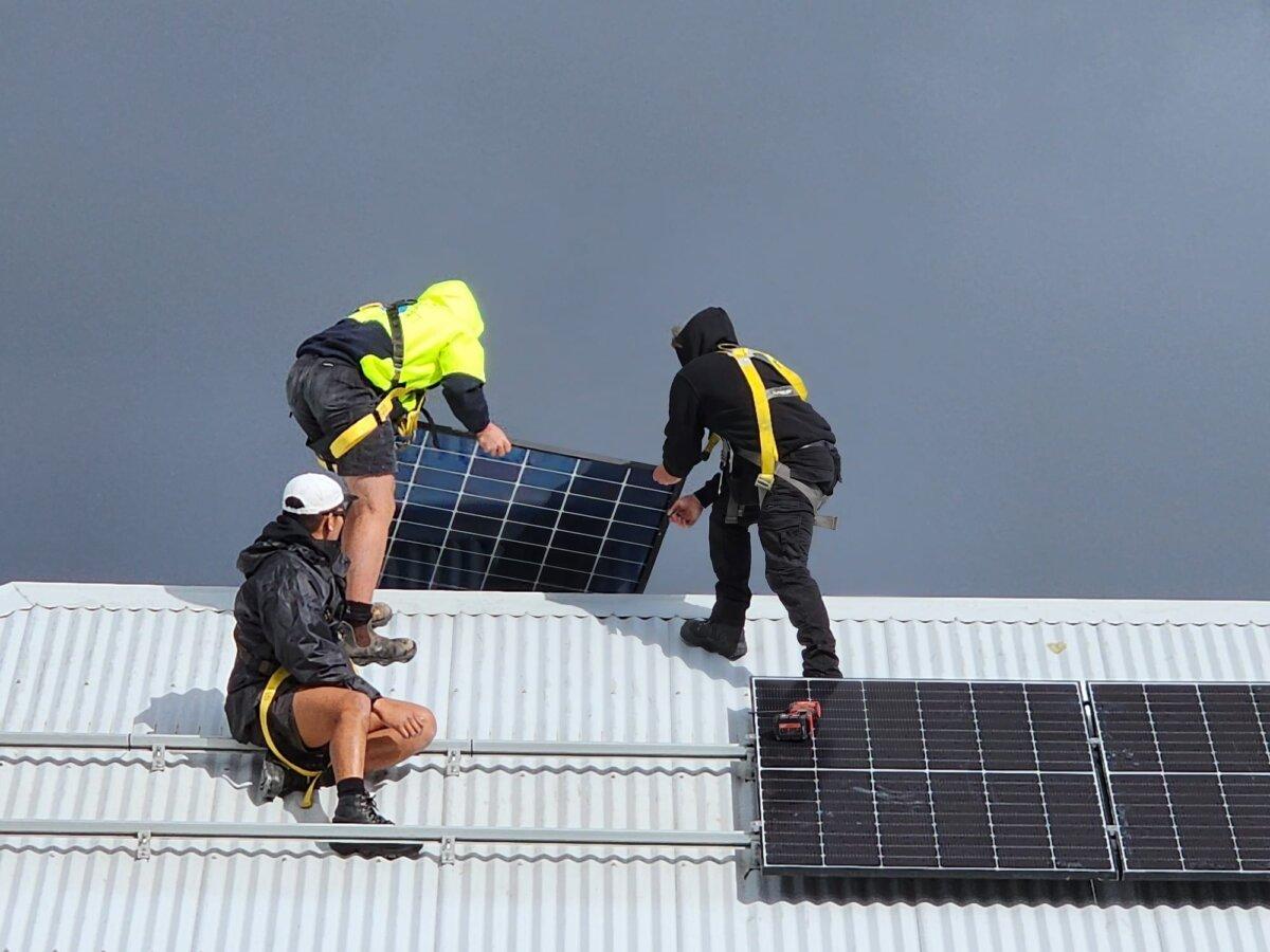 Three men install solar panels on a roof as storm clouds approach in Albany, Western Australia, on Aug. 29, 2024. (Susan Mortimer/The Epoch Times)