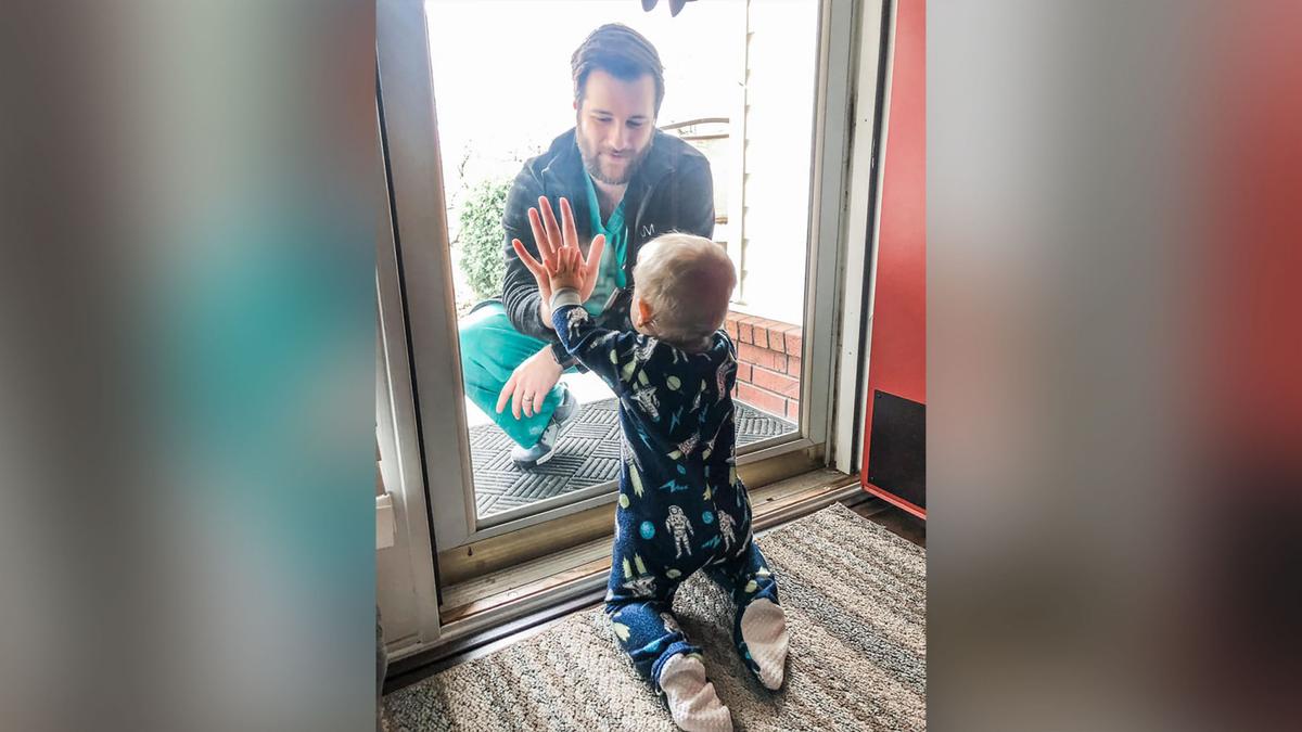 Dr. Jared Burks sees his 1-year-old son crawl for the first time, as he touches a glass door from the outside while their son Zeke touches it from the inside of their Jonesboro, Ark. home in this photo posted on Facebook, on March 25, 2020. (Burks Family via AP)