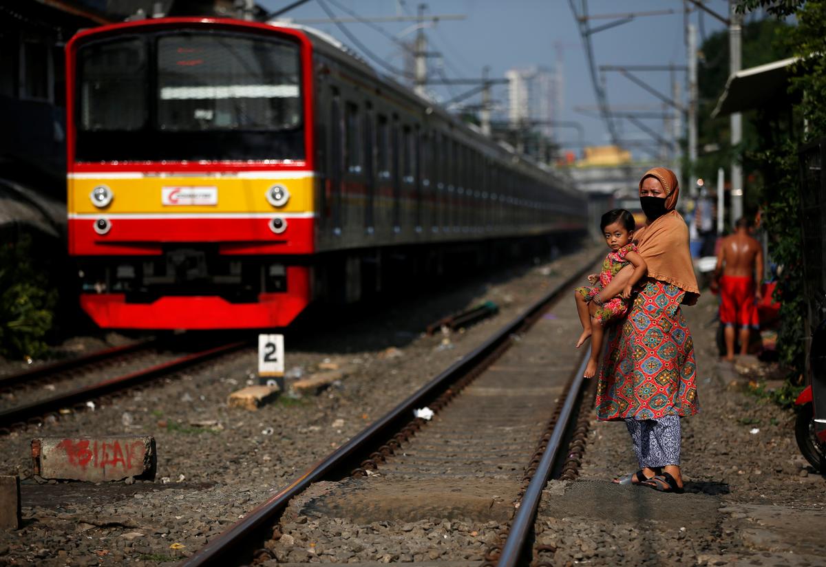 A train passes by a woman wearing a protective mask and holding a child along rail tracks in Jakarta, Indonesia, on April 12, 2020. (Willy Kurniawan/Reuters)