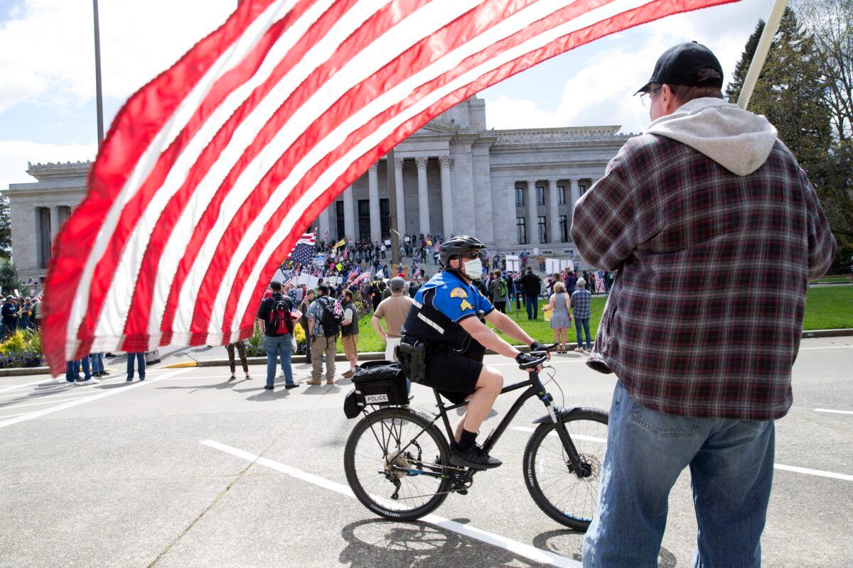 A law enforcement officer wearing a mask rides a bike past a demonstrator with a flag during a 'Hazardous Liberty! Defend the Constitution!' rally at the Capitol building in Olympia, Washington on April 19, 2020. (Karen Ducey/Getty Images)