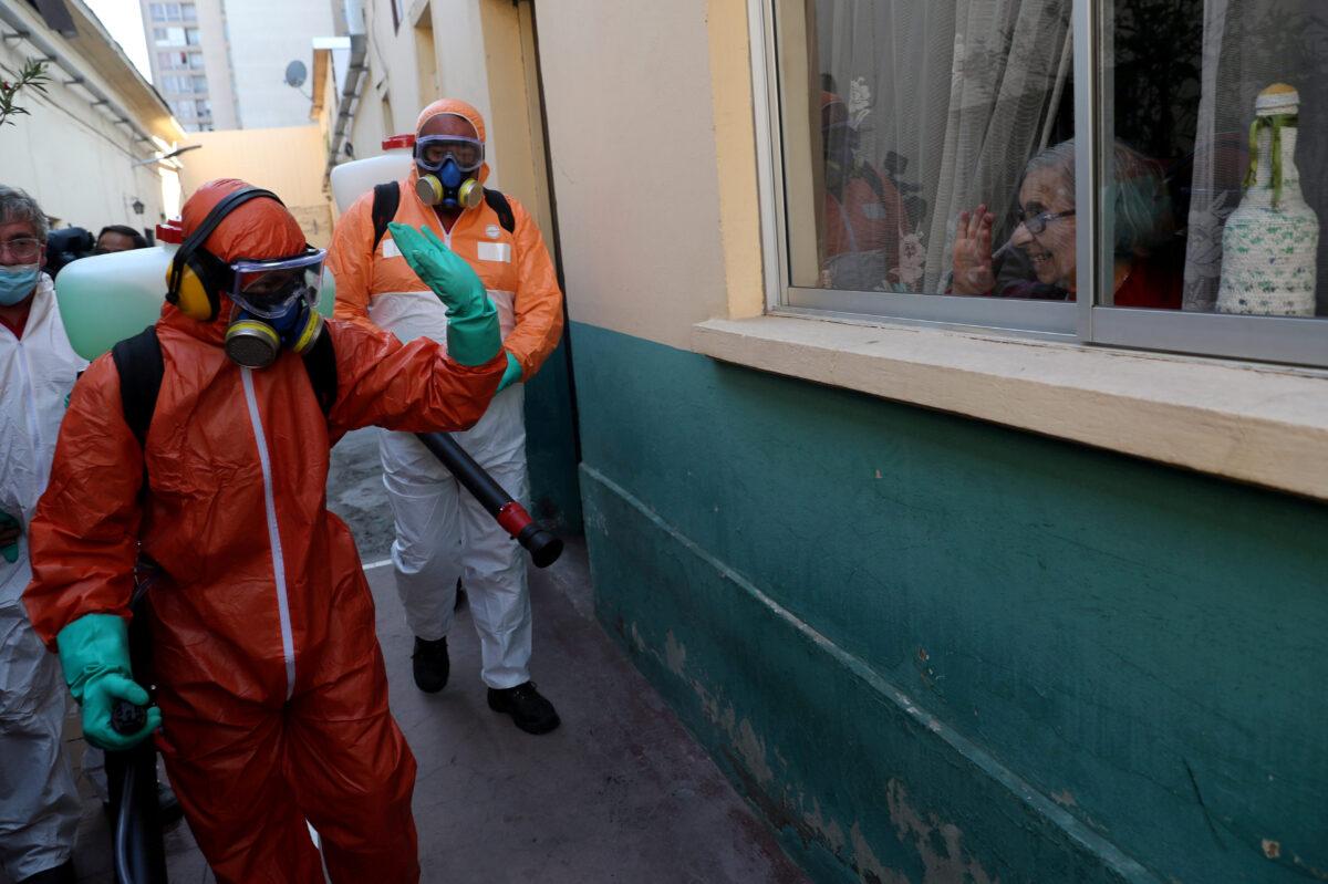 A worker wearing protective gear waves to a woman while using disinfectant to clean outside of the residences, following the CCP virus outbreak in Santiago, Chile on April 15, 2020. (Ivan Alvarado/Reuters)