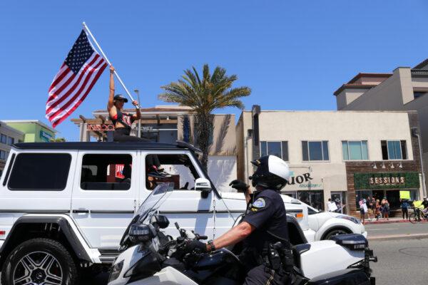 A motorcycle policeman tells a woman with a flag to go back inside her vehicle at a protest gathering in Huntington Beach, Calif., on May 1, 2020. (Jamie Joseph/The Epoch Times)