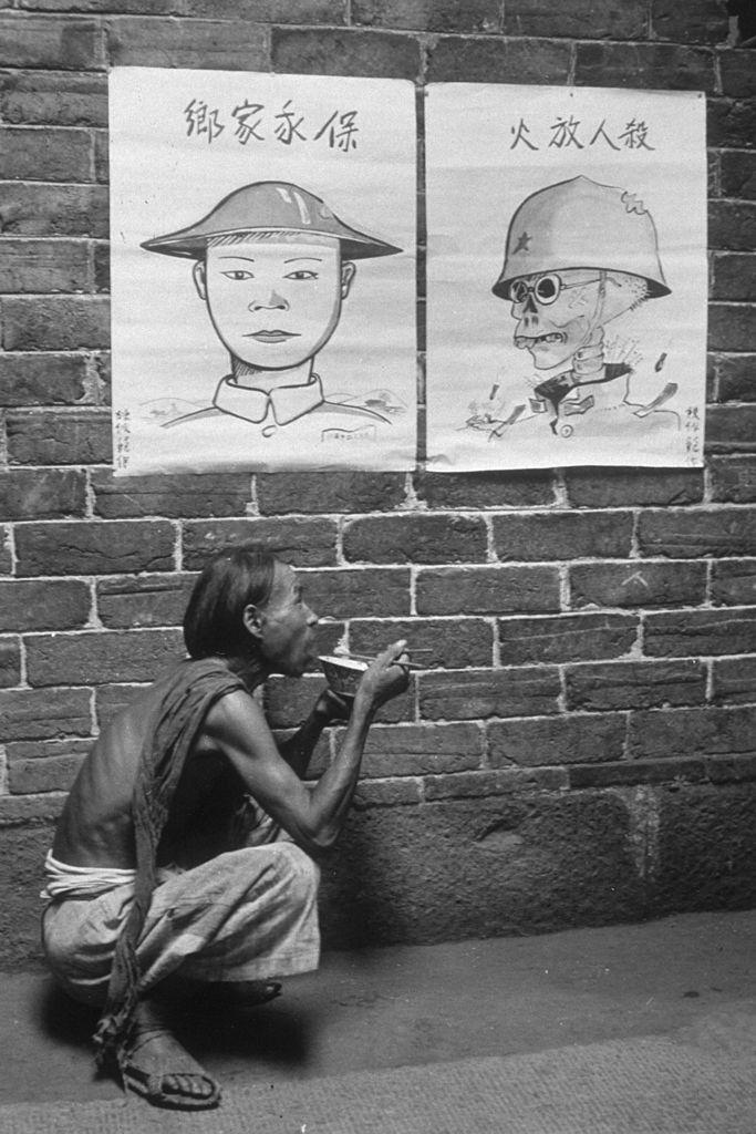 A man with his ribs showing eats rice squatting in front of Chinese government propaganda posters during the Sino-Japanese War. (Hulton Archive/Getty Images)
