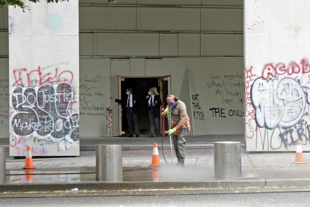 A worker washes graffiti off the sidewalk in front of the Mark O. Hatfield Federal Courthouse in downtown Portland, Ore., as two agents with the U.S. Marshals Service emerge from the boarded-up main entrance to examine the damage, on July 8, 2020. (Gillian Flaccus/AP Photo)