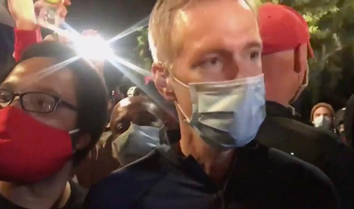 In this still image from video, Portland Mayor Ted Wheeler, right, stands at a fence guarding a federal courthouse as tear gas drifts by, in Portland Ore., on July 23, 2020. (Karina Brown via AP)