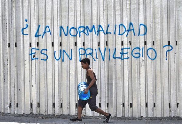 A boy carries a drum with water as he walks past a graffiti reading “Is Normality a Privilege?” during a new power outage in Venezuela, at Fuerzas Armadas Avenue in Caracas on March 31, 2019. (FEDERICO PARRA/AFP via Getty Images)