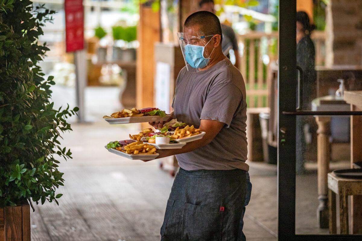 A waiter wearing protective face gear balances plates of food on his arms as he carries them to diners at a restaurant in Newport Beach, Calif., on Sept. 9, 2020. (John Fredricks/The Epoch Times)