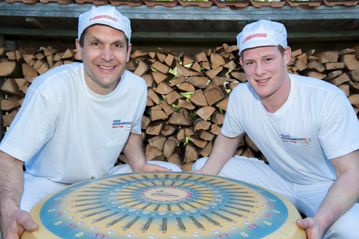 Cheesemakers Marcel Zuger and Robin Straub holding a 200-pound wheel of Emmentaler. (Courtesy of Gourmino)
