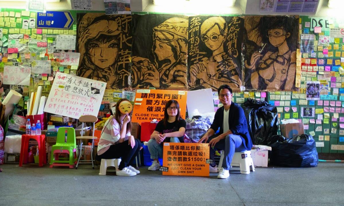 Three people sit in front of a Lennon Wall displaying hand-painted posters by Otto Yuen, in Hong Kong. (Courtesy of Otto Yuen)