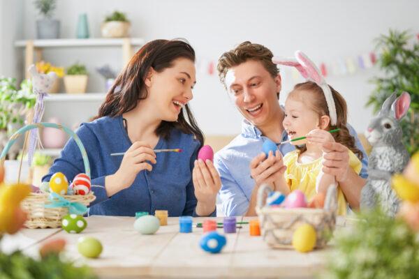 Mother, father and daughter are painting eggs. Happy family are preparing for Easter. Cute little child girl wearing bunny ears. (Shutterstock)