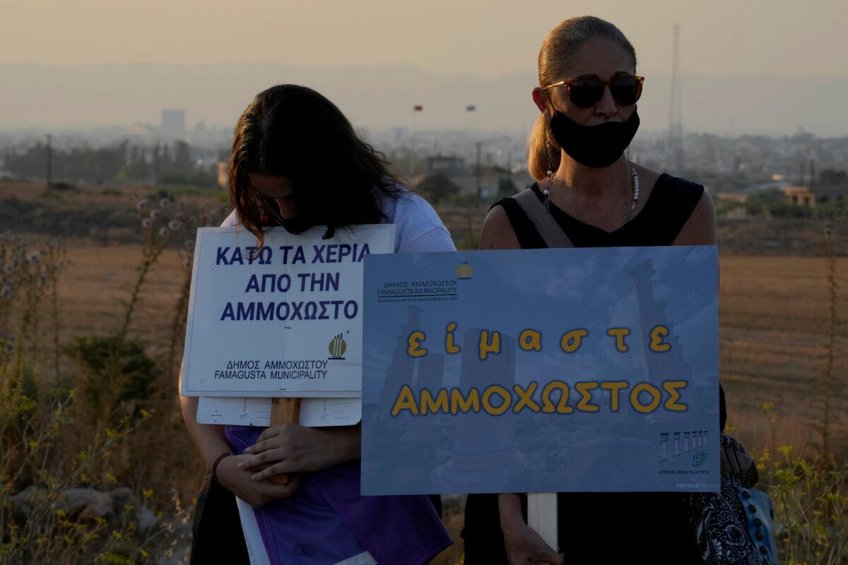 Residents of Varosha or Famagusta, abandoned city, in the background, hold placards that read in Greek: 'We are Ammochostos," right, and "Hands down of Famagusta" during a protest against the Turkish President visiting the Turkish occupied part of the island at the north and the 47th anniversary against the Turkish invasion in the island, in Dherynia, Cyprus, on July 19, 2021. (Petros Karadjias/AP Photo)