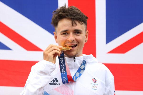 Thomas Pidcock of Team Great Britain bites his gold medal and pose with the flag of his country in the background after the Men's Cross-country race on day three of the Tokyo 2020 Olympic Games at Izu Mountain Bike Course in Izu, Shizuoka, Japan, on July 26, 2021. (Michael Steele/Getty Images)