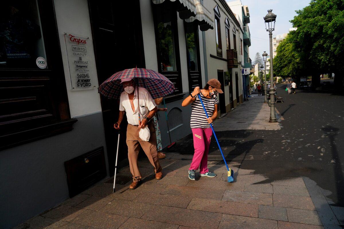 A man walks with an umbrella as women clean the ash from the street following the eruption of a volcano on the Canary Island of La Palma, in Tazacorte, Spain, on Oct. 1, 2021. (Juan Medina/Reuters)
