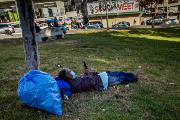 A homeless man wearing a facemask lies on the grass while watching his phone in MacArthur Park in Los Angeles on May 21, 2020. (Apu Gomes/AFP via Getty Images)