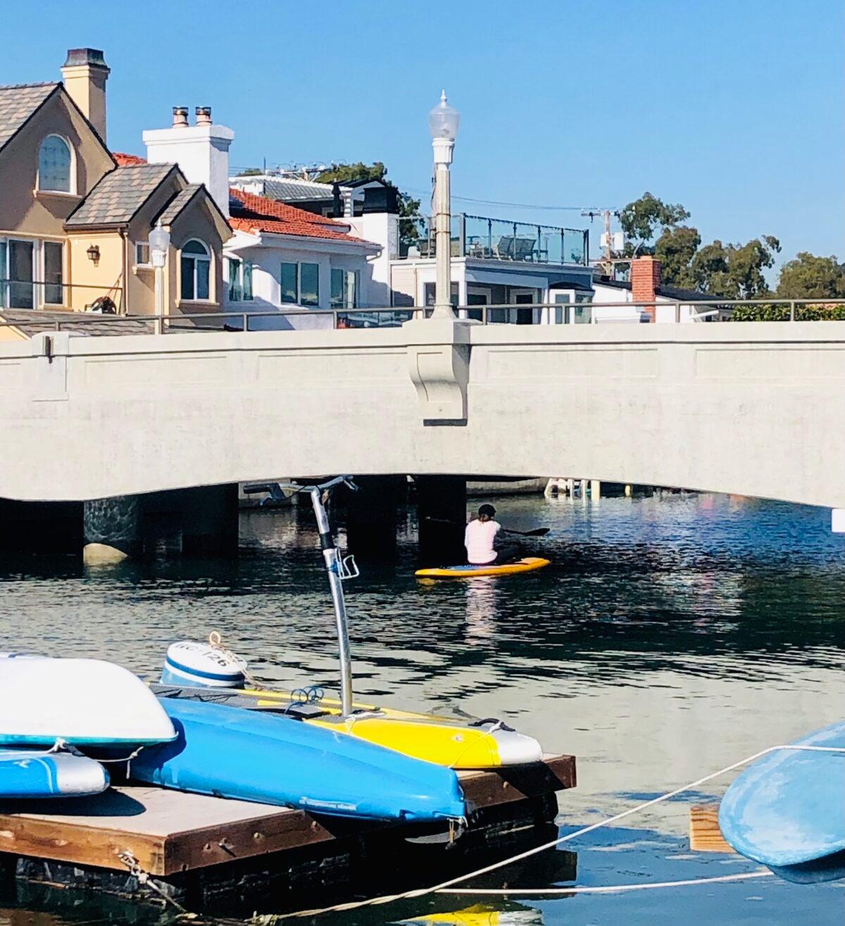 A paddle boarder has to sit to go under a bridge during king tides in Newport Beach, Calif., on Jan. 4, 2022. (Lynn Hackman/The Epoch Times)