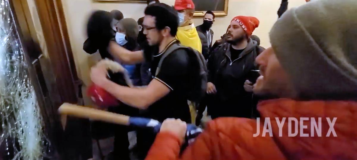 Chad Barrett Jones (in the red jacket and gray cap) uses a wooden flagpole to break a window outside the Speaker's Lobby at the U.S. Capitol on Jan. 6, 2021. (Video Still/Jayden X)
