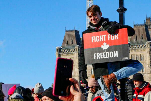 A protester climbs a lamppost to display his sign on Parliament Hill in Ottawa on Jan. 29, 2022. (Noé Chartier/The Epoch Times)