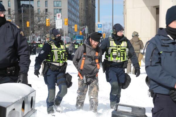 Police walk a detained person away outside the Westin Hotel in Ottawa on Feb. 18, 2022. To the rear, the protest heats up, leading to many arrests. (Richard Moore/The Epoch Times)