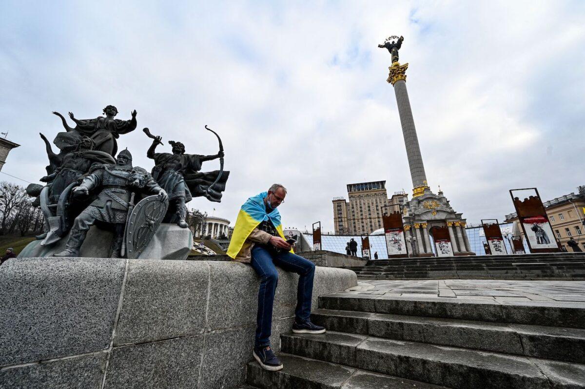 A man wrapped with a Ukrainian national flag watches news on his mobile phone as he sits at Maidan Independence Square in Kyiv on Feb. 24, 2022, the day that Russian ground forces invaded Ukraine. (Sergei Supinsky/AFP via Getty Images)