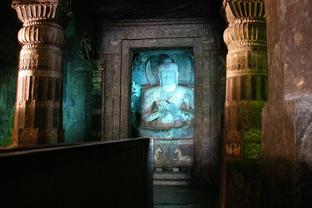 Surreal lights illuminate a shrine for Buddha in Cave 17. The shrine has an ornately carved door frame with sections that depict floral designs, various Buddha figures, and other decorative elements. Murals and ornate columns are also features of Cave 17. (Vyacheslav Argenberg/ CC BY-SA 4.0)