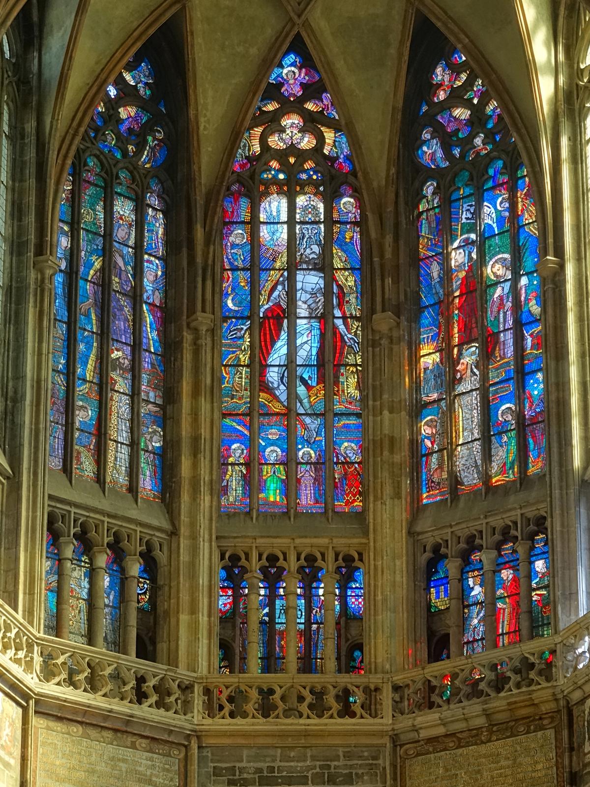 In this triptych, the stained glass windows with angled sides appear on the south side of the cathedral. Their glowing quality creates a sense of awe. In the center, Jesus is held in the hands of the Creator, and above a white dove appears, symbolic of the Holy Trinity. On the left, we see the Virgin Mary and, on the right, St. Wenceslas and St. Vitus on their knees. (ErwinMeier/CC BY-SA 4.0)