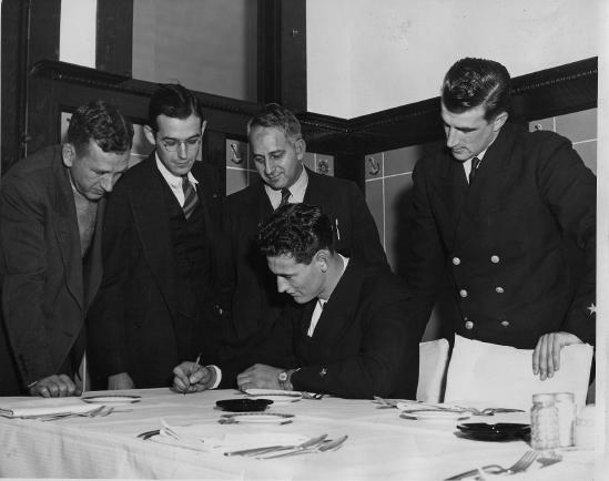 Ted Williams in his<br/>U.S. Navy uniform signing autographs in Kokomo, Indiana in 1944. Others in the photograph (L-R): Ralph King, Richard “Dick” Rainbolt, Haines Sleeth, Johnny Pesky. Pesky was Williams’s teammate in Boston. (Public Domain)
