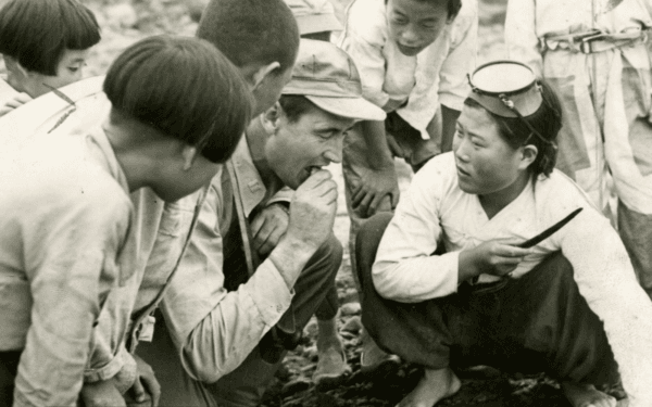 While searching for photographic subjects in a remote Korean fishing village in June<br/>1953, Ted Williams paused to eat a live clam offered by a native diving woman. Korean children watch his reaction. (Public Domain)
