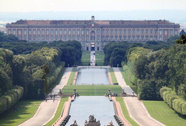 Standing five stories tall and 810 feet wide, the Royal Palace of Caserta is the largest palace by volume in the world. This view of the back side of the palace only hints at the incredible two-mile-long promenade and waterway that stretch vastly across the Italian landscape like a man-made river adorned by impressive fountains and sculptures that are framed by natural forests. (<a class="external text" href="https://web.archive.org/web/20161029173241/http://www.panoramio.com/user/410152?with_photo_id=24347479" rel="nofollow">Carlo Pelagalli</a>/<a class="mw-mmv-license" href="https://creativecommons.org/licenses/by-sa/3.0" target="_blank" rel="noopener">CC BY-SA 3.0</a>)