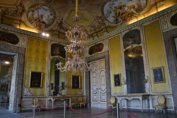 An example of the Baroque decorative rooms at the Royal Palace with all of its myriad details and excessive ornamentation. The entire ceiling is elaborately painted with elements of illusory dimension and windows. Gold-painted borders stretch from floor to ceiling outlining the brightly colored walls and large mirrors and doors. A gaudy, glass chandelier hangs low with multiple colors fighting for the viewer's attention in this richly Baroque room. (Carlo Dell'Orto/CC BY-SA 4.0)