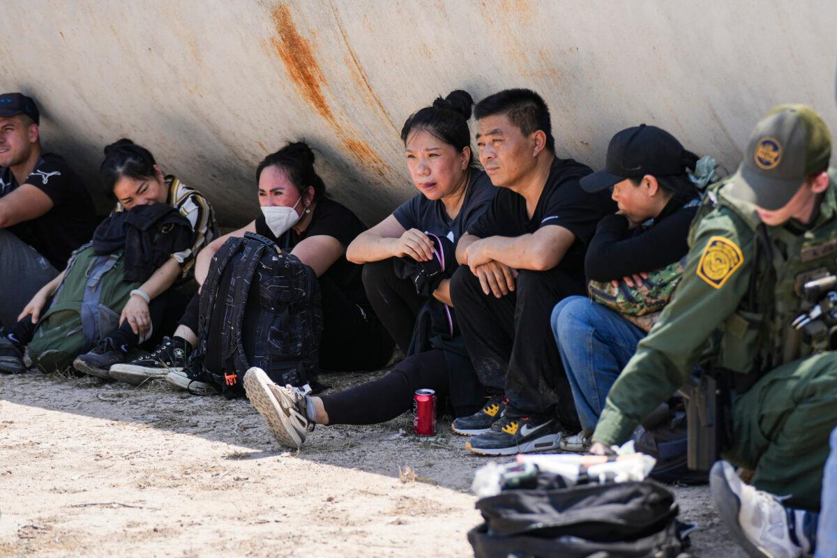 Five Chinese illegal immigrants wait while a Border Patrol agents provides medical care to a woman within a large group of illegal immigrants near Eagle Pass, Texas, on May 20, 2022. (Charlotte Cuthbertson/The Epoch Times)