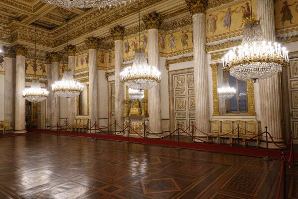 The ballroom of the Royal Palace of Turin is an opulent space lit by eight shining chandeliers. A richly-detailed lacunar, a ceiling constructed with recessed panels, creates depth and complexity which is reflected by simulated coffer designs on the floor below. Large white pillars reach from floor to ceiling and a continuous mural of dancing women runs along the top of the walls below gold-painted cornices and bronze capitals. (Guilhem Vellut/CC BY 2.0)