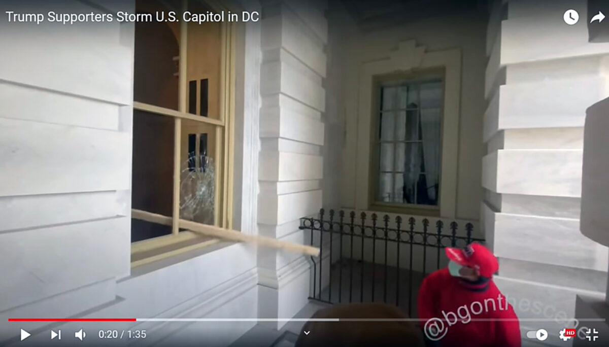 A man known only by the hashtag #RedOnRedGlasses throws a 2-by-4 through a Capitol window on Jan. 6, 2021. (Sedition Hunters/Screenshot via The Epoch Times)