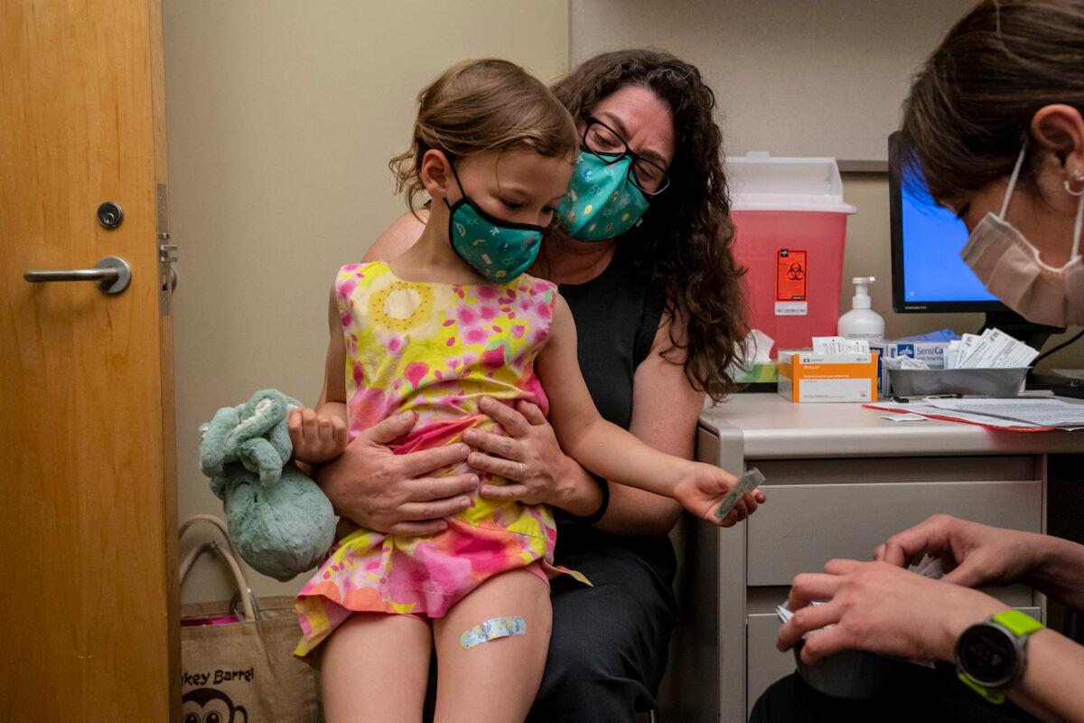 A 3-year-old girl sits on the lap of her mother and gets a sticker from the nurse after receiving her first dose of the Pfizer COVID-19 vaccine at UW Medical Center–Roosevelt in Seattle, Washington, on June 21, 2022. (David Ryder/Getty Images)