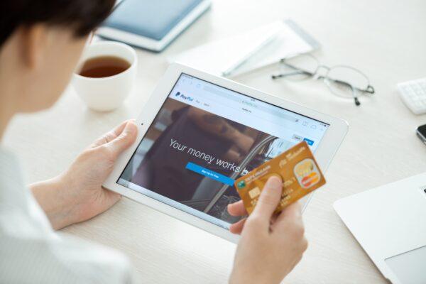A person holds a credit card, looking at an iPad with the Paypal website on the screen. (Antlii/Shutterstock)