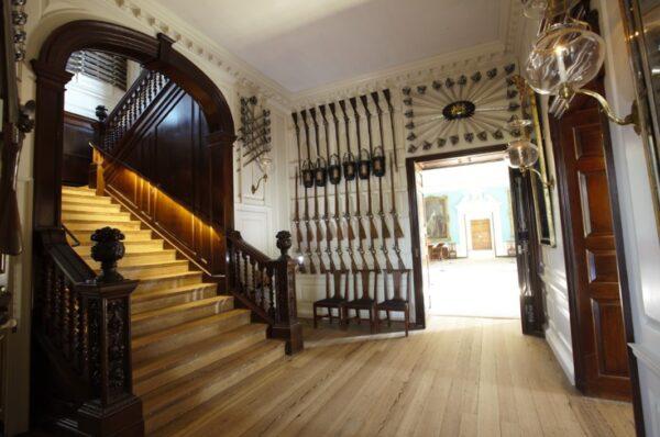 The display of guns and swords continue to decorate the walls in the first floor hallway and up the stairs. Walls of American Walnut give the room a reddish tone, while the pine floors lighten the effect. (J.H. Smith/Cartio)