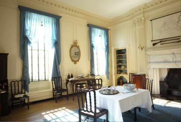 Social engagements and other business would also occur in the first floor dining room. George Washington was a frequent visitor. The wood paneling, cornice and pilasters, and flattened columns are painted in a neutral cream. Curtains in a muted blue grace large windows that reach the ceiling. The mantel and mantel shelf of Italian marble in a delicate mauve frames the fireplace. Built-in shelves in a corner display items needed for the dining room. (J.H. Smith/Cartio)