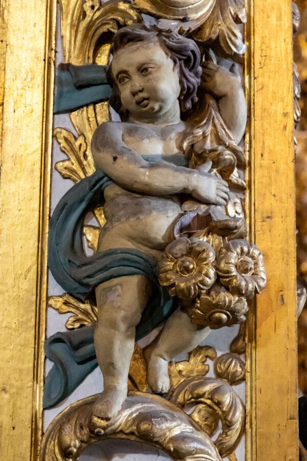 One of the details of the church’s entrance hall is a bas-relief of a putto holding flowers, in an elaborate framing. Putti are little boy figures, sometimes with wings, that are often depicted in Baroque and Rococo art. The gilded woodwork combines Portuguese and Brazilian artistry. (<a href="https://en.wikisource.org/wiki/Author:Paul_Robert_Burley">Paul R. Burley</a>/ <a href="https://creativecommons.org/licenses/by-sa/4.0/deed.en">CC BY-SA 4.0</a>)