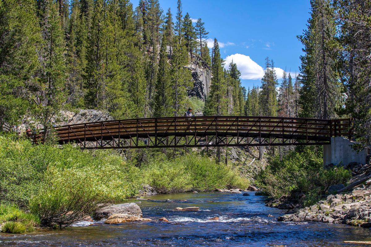 A visitor walks across a bridge over San Joaquin Creek at Soda Springs Meadow, with a view of Devils Postpile National Monument in the background, near Red's Meadow Resort & Pack Station on June 21, 2022, in Mammoth Lakes, California. (Allen J. Schaben/Los Angeles Times/TNS)