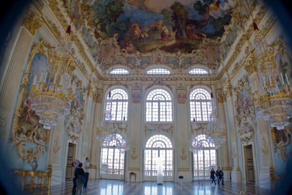 The great hall, at the center of the palace, is a striking two-story room filled with light. The stucco decoration, ceiling fresco, and gold details are in the ornamental Rococo style, designed by 18th-century Bavarian artist Johan Baptist Zimmermann. The room has soft pastel colors on the walls and a colorful ceiling depicting the nymph and flower goddess Flora, who brings nature into the palace. This idyllic scenery honors the prosperity of the state of Bavaria. (<a href="https://upload.wikimedia.org/wikipedia/commons/a/a1/Steinerner_Saal%2C_Palacio_de_Nymphenburg%2C_M%C3%BAnich%2C_Alemania14.JPG">Diego Delso/CC BY-SA 3.0</a>)<span style="font-size: 16px;"> </span>