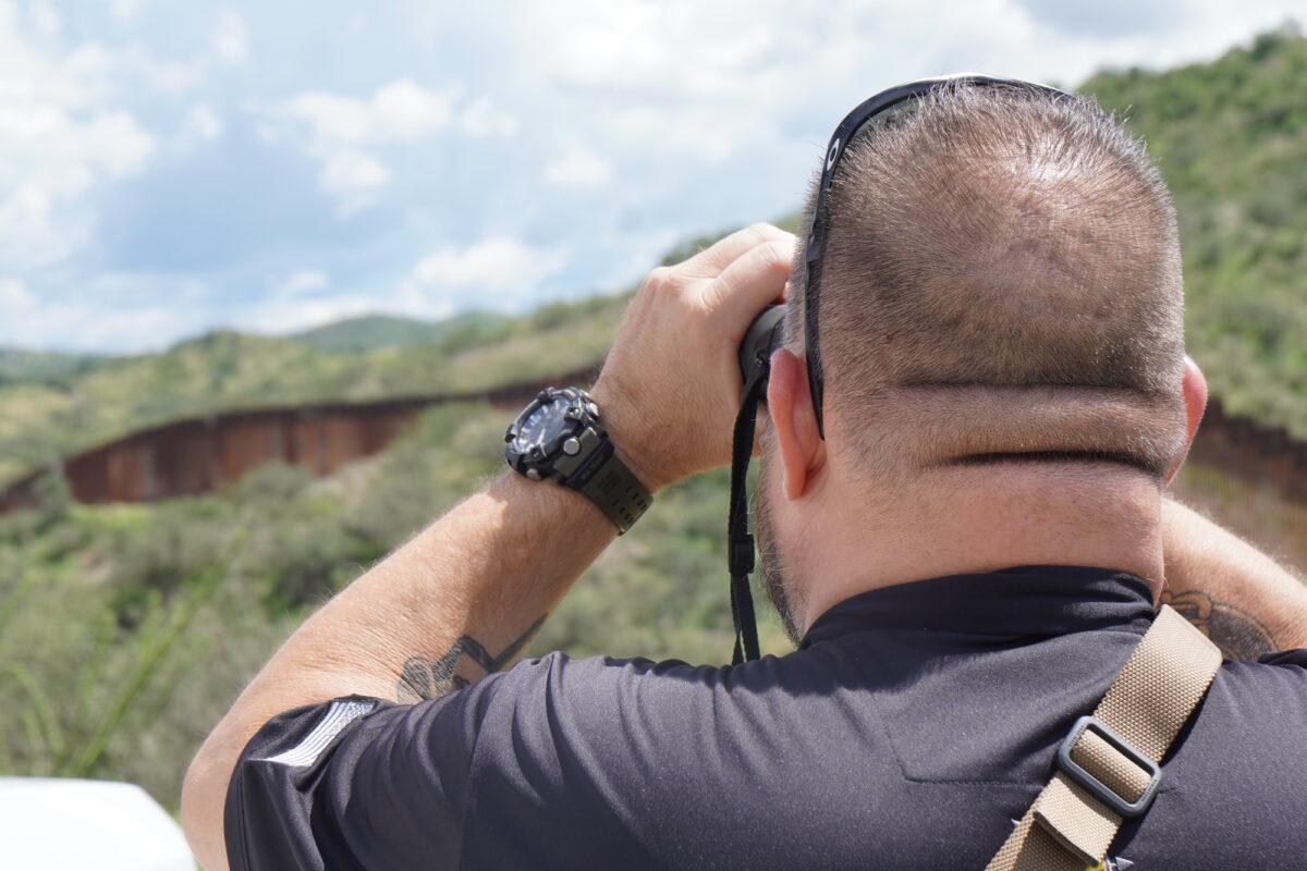 Sam, the owner of a private security firm in Arizona, keeps watch with binoculars over the U.S. border wall with Mexico on Aug. 25. (Allan Stein/The Epoch Times)
