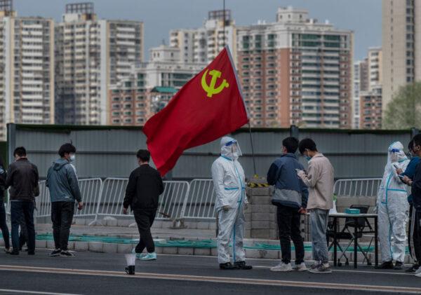 A Communist Party of China flag is seen next to a health worker wearing protective clothing as office workers line up for nucleic acid tests to detect COVID-19 at a makeshift testing site on April 28, 2022 in Beijing, China. (Kevin Frayer/Getty Images)