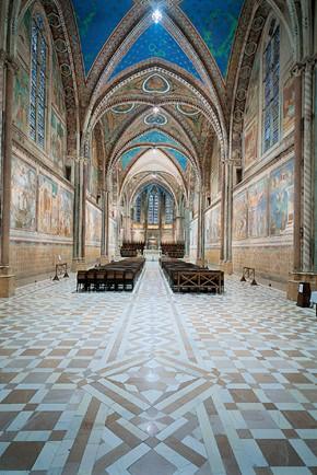 The strikingly colorful, airy, and majestic nave of the upper church constructed as a simple, single four-bay nave with a cross-vaulted ceiling, and floor patterns of leaves and crosses. Golden stars decorate four ribbed vaults on the blue ceiling. Unlike the design of the lower basilica, the clustered columns have ribs in a Gothic style. Tall Gothic stained-glass windows line the nave. The frescos along the walls depict scenes by artists Pietro Cavallini, Cimabue, and Jacopo Torriti. (Public Domain)