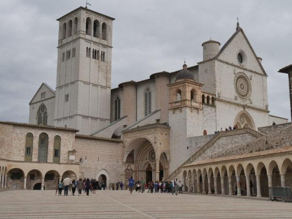 The lower and upper churches of the basilica are seen from the Lower Plaza of St. Francis. The façade of the basilica uses the French Gothic style with white-washed brick, a split doorway, and a heavy bell tower. The entrance is seen in the center of the building under a large arch and is divided by a column. On the right, the friary of St. Francis is composed of 53 Romanesque-style arches, supported by buttresses. Today, the friary behind the arched colonnade houses a library and a museum holding pilgrims’ donations. (<a class="jss337 jss113 jss115 jss114" href="https://www.shutterstock.com/image-photo/assisi-external-st-francis-basilica-one-1273488544">FilippoPH</a>/Shutterstock)
