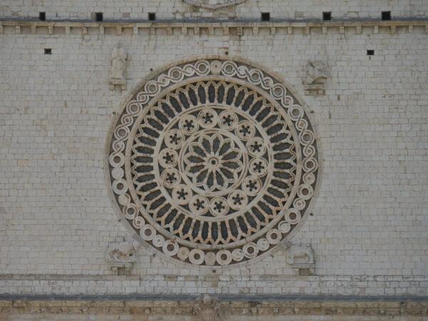 In the tympanum (a decorative wall surface above an entrance, door, or window) above the two cusped arches of the basilica’s exterior, there is a rose window with carvings, which are typical Romanesque decorative details. It is enclosed in a Renaissance-style porch with large white stone walls and arched windows. This window is often called the “eye of God” or “the eye of the most beautiful church in the world,” according to author Gualtiero Belucci in “Assisi, Heart of the World." (<a href="https://www.shutterstock.com/image-photo/assisi-rosette-st-francis-basilica-1273490557">FillipoPH/Shutterstock</a>)