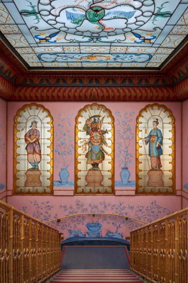 The colorful entrance hall is in the Regency Chinoiserie style, inspired by Chinese décor with gilding, lacquering, and oriental figures. Other Chinese features include the bamboo motif, the blueish-green birds on the pink wallpaper, and the bamboo staircase at the end of the hall. There are more Chinese elements such as figurines and vases, silk tassels, and hexagonal lanterns. The hall is arranged symmetrically, with Gothic elements that include the carved wood furniture and the stained-glass windows. (Brighton & Hove Museums)