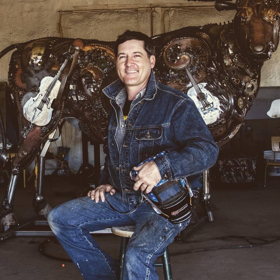 John Lopez in the studio with a scrap metal cow in the background. (Courtesy of <a href="https://www.instagram.com/johnlopezstudio/">John Lopez</a>)