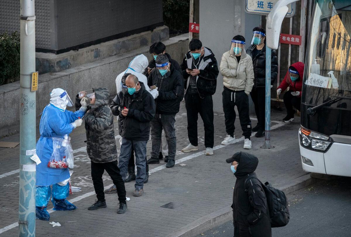An epidemic control worker wears PPE to prevent the spread of COVID-19 as he gives a nucleic acid test to a resident as others wait in line wearing face shields before leaving by bus from an area with communities in lockdown in Beijing, China, on Nov. 30, 2022. (Kevin Frayer/Getty Images)
