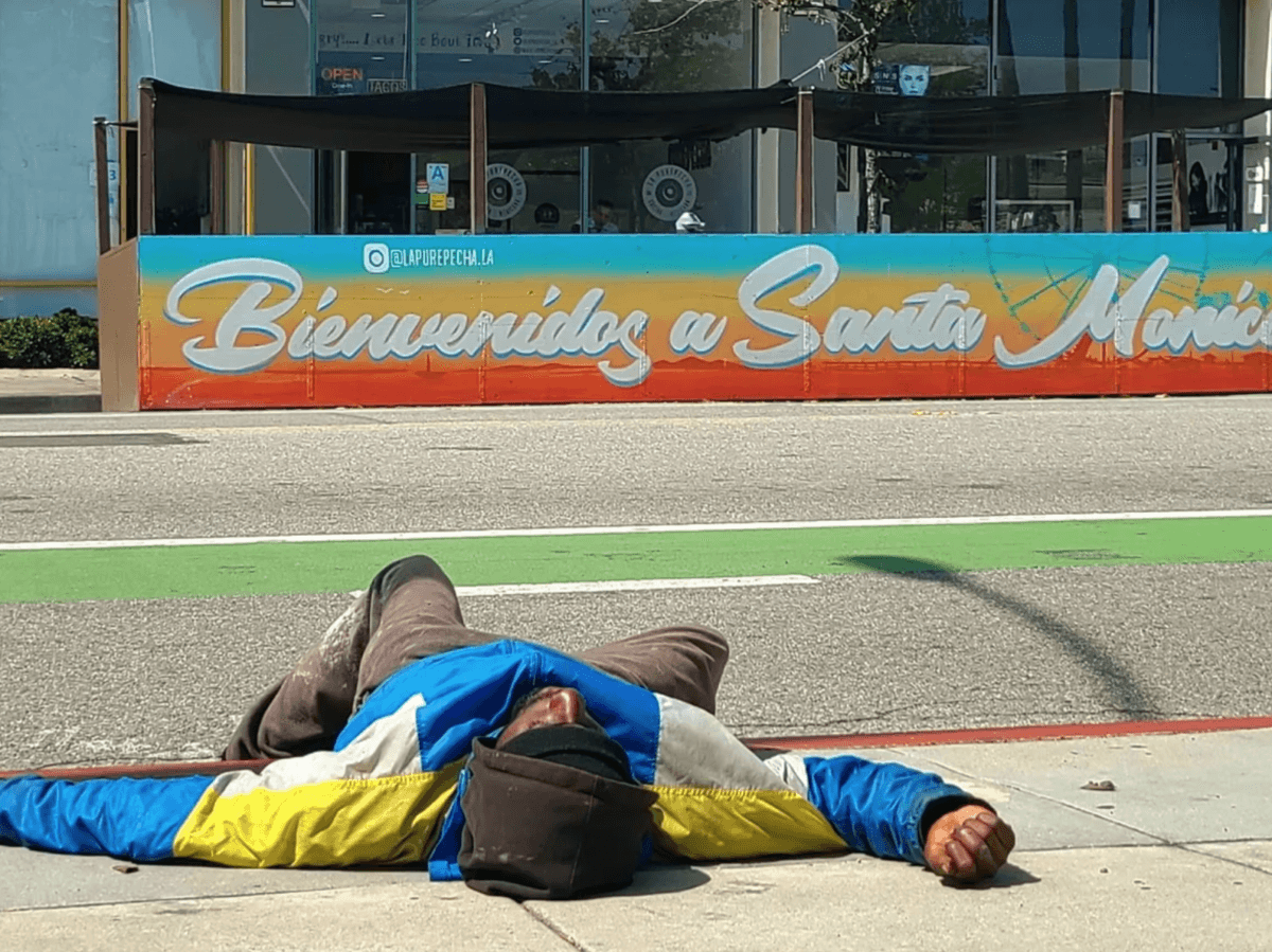 A homeless man lays on the sidewalk across from a sign saying "Welcome to Santa Monica" in Santa Monica, Calif. (Courtesy of The Santa Monica Coalition)