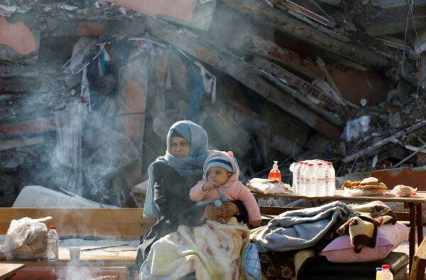 A woman holding a child sits by a collapsed building as search for survivors continues, in the aftermath of a deadly earthquake in Hatay, Turkey, on Feb. 10, 2023. (Umit Bektas/Reuters)